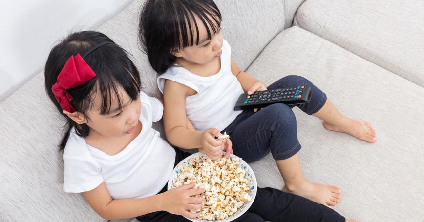 Two girls on a couch watching TV while eating popcorn