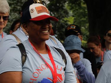 A Black female veteran in a blue t-shirt and American flag hat smiles while standing outside during an all female veteran honor flight