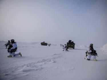 U.S. soldiers kneel on the ground in an open, snowy landscape keeping watch. A couple armored vehicles are in the background.