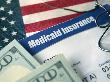 Closeup of Medicaid card on top of a U.S. flag with 100-dollar bills and a stethoscope on top, photo by Kameleon007/Getty Images