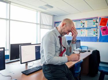 Stressed-looking male teacher leaning against a desk with one hand on his forehead, photo by SolStock/Getty Images