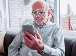 Senior man holds smart phone and it performs a scan of his face, photo by mikkelwilliam/Getty Images