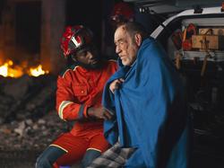 African American man in paramedic uniform asking questions to senior man wrapped in blanket