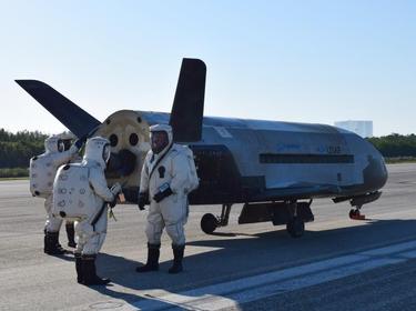 The experimental X-37B space plane surrounded by three  personnel in sealed suits